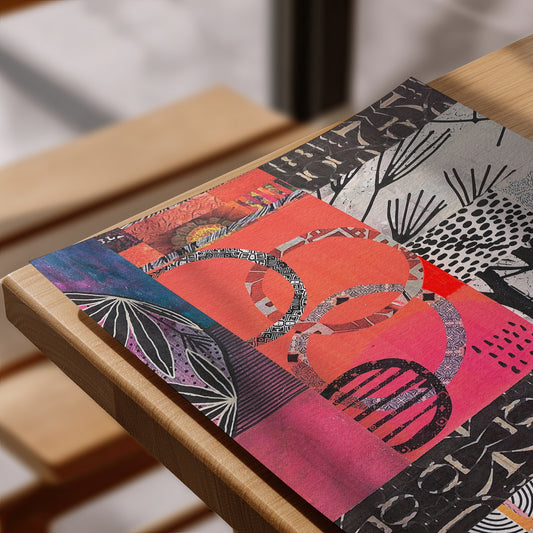 A close-up of a colorful abstract art print or poster laid diagonally on a light wood table. The artwork features a patchwork of pink, coral, purple, black, and white patterns, including leaf motifs and geometric shapes.