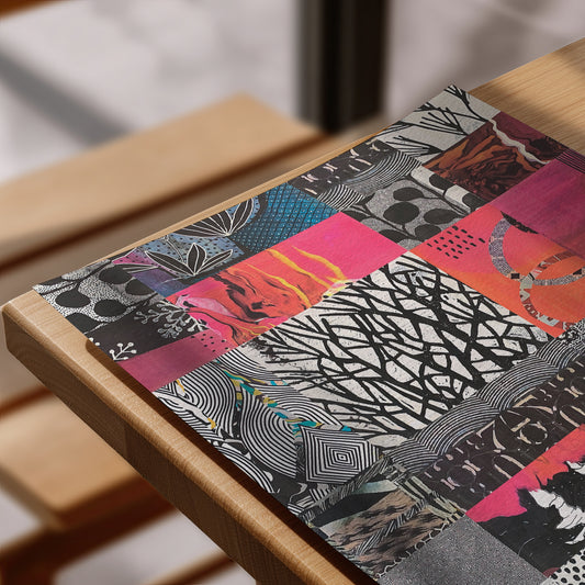 A close-up of a colorful patchwork placemat featuring various abstract patterns and vibrant pink, orange, blue, and black sections, laid on a light wood table.