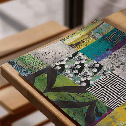 A rectangular art print with a vibrant patchwork design featuring green leaves, black-and-white flowers, geometric patterns, and script text, displayed on a light wood frame.