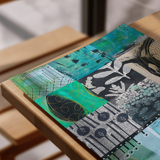 A rectangular mixed-media art print or placemat featuring an abstract collage of teal, green, black, white, and gray textures, shown resting on a light wood table.
