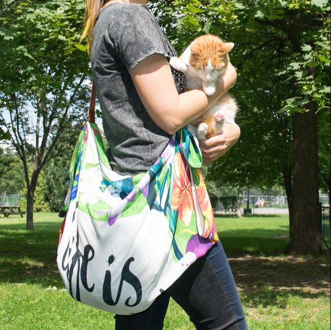 Person holding a colorful cat and a floral-patterned bag with 'life is' text in a park setting.