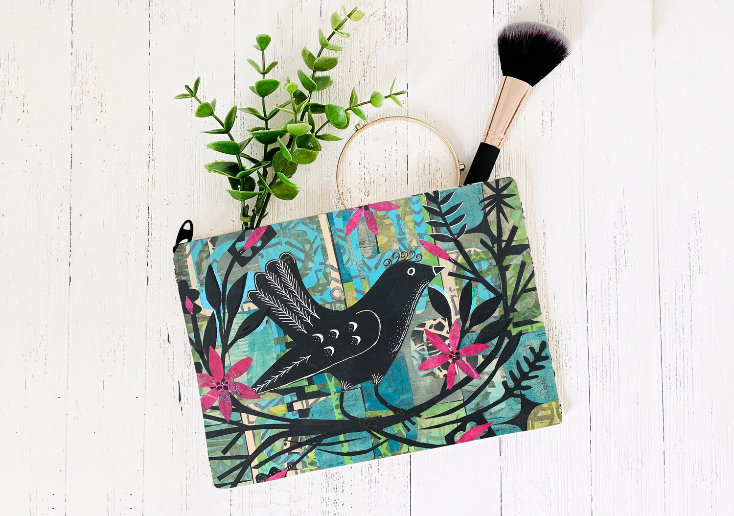 Colorful pouch with bird design on a white wooden surface with a plant and makeup brush.