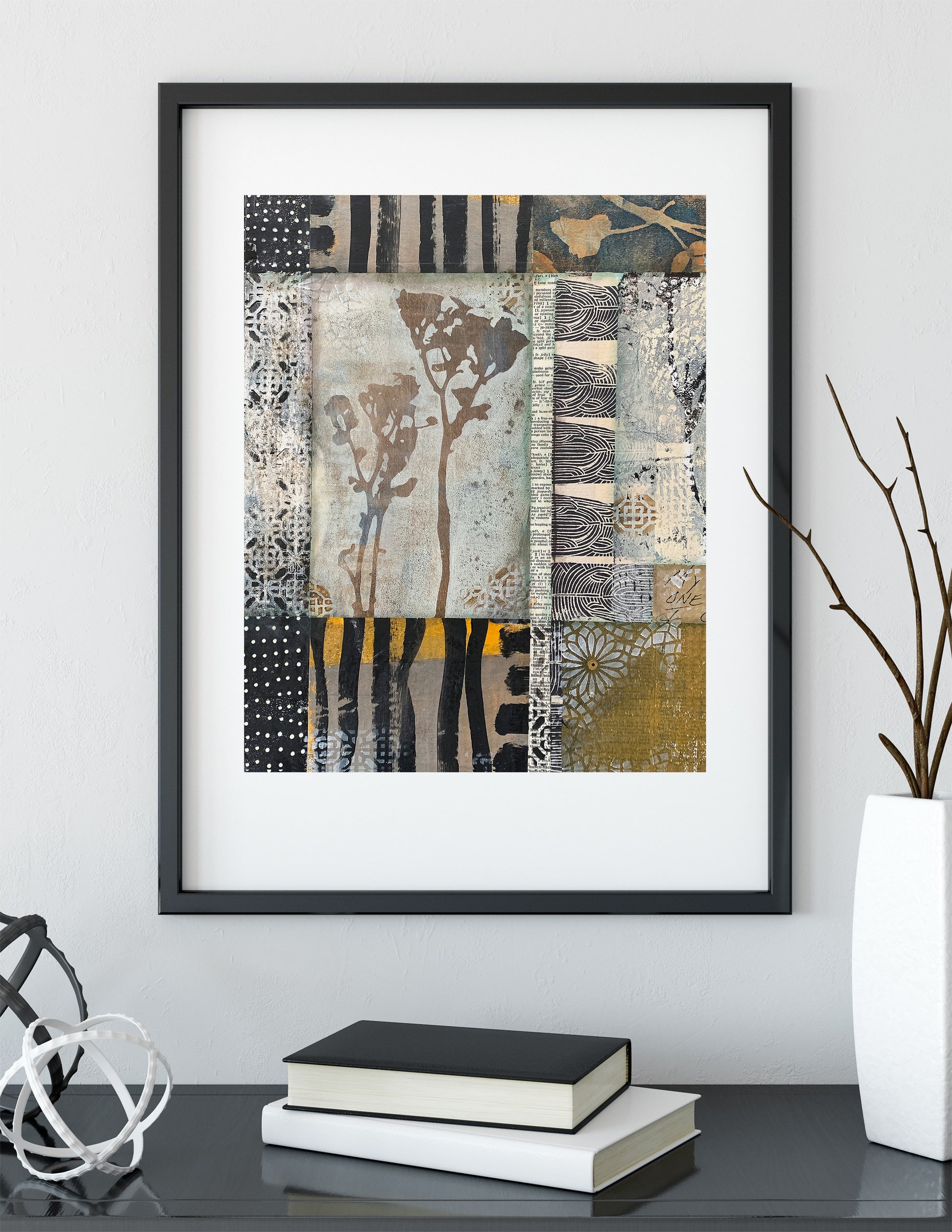 Framed abstract wall art with a patchwork textile-like design in black, beige, gray, white, and muted yellow, displayed above a table with books and decorative objects in a styled interior setting.