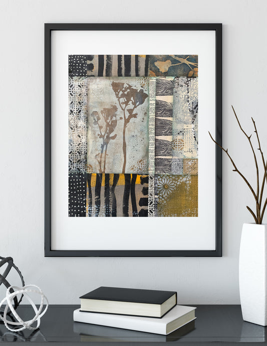 Framed abstract wall art with a patchwork textile-like design in black, beige, gray, white, and muted yellow, displayed above a table with books and decorative objects in a styled interior setting.