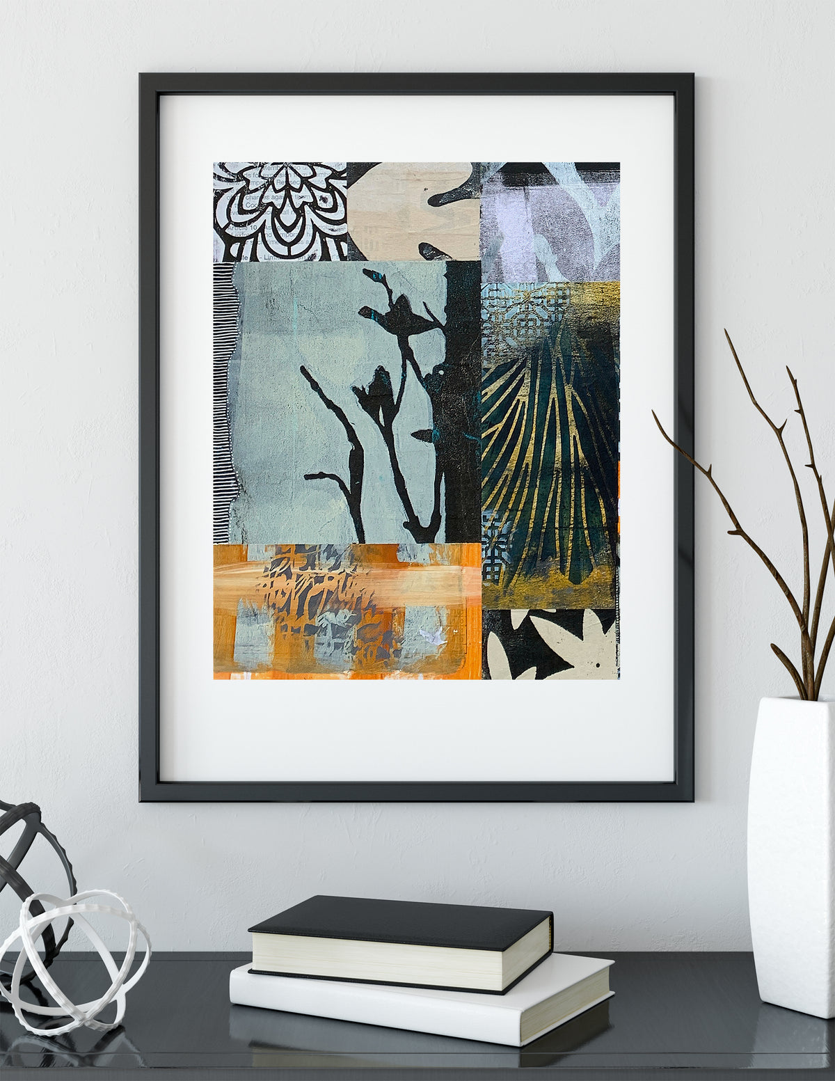 A framed abstract art print with a black frame and white mat, hanging on a light gray wall above a dark table with books and decorative objects.