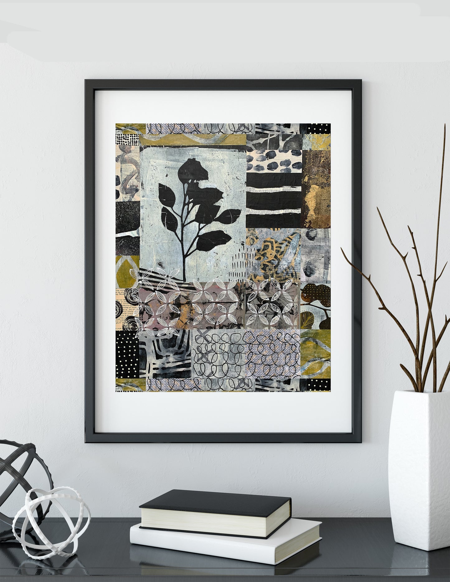 A framed abstract art print featuring a collage of black, white, gray, and olive green patterns with a central black rose motif, displayed on a shelf in a modern interior setting.