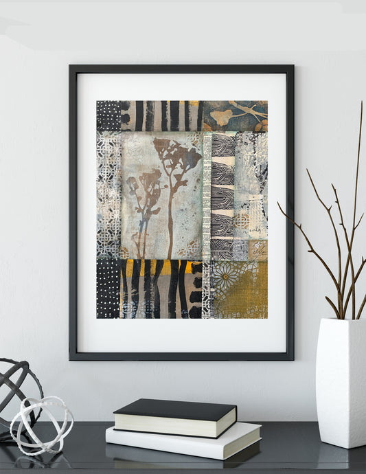 A framed abstract art print with a black frame and white mat, featuring a patchwork design in black, beige, gray, and gold tones. The framed print is displayed on a dark table against a light gray wall, styled with books and a white vase with branches.
