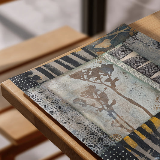 A rectangular placemat with an abstract patchwork design featuring tree silhouettes, stripes, and textured patterns in muted blue, beige, gray, black, and yellow, displayed on a light wood table.