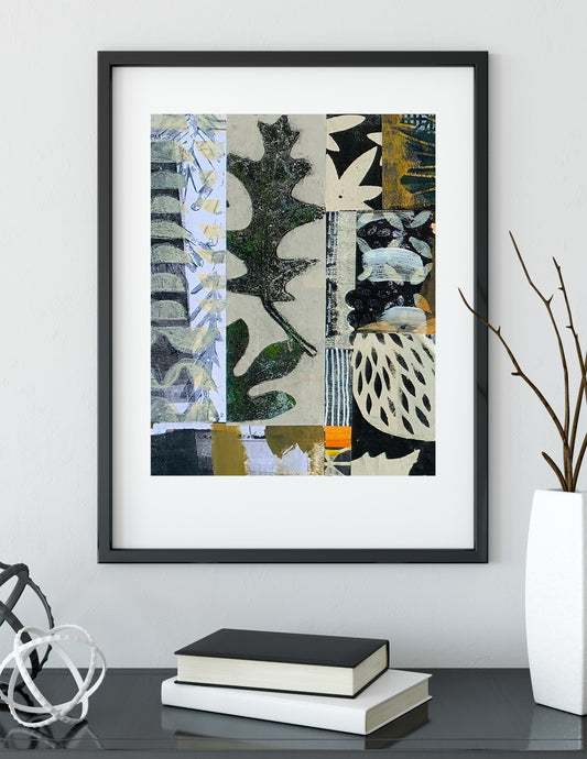 A framed abstract art print with a black frame and white mat, displayed on a dark table with books and a white vase against a light gray wall.