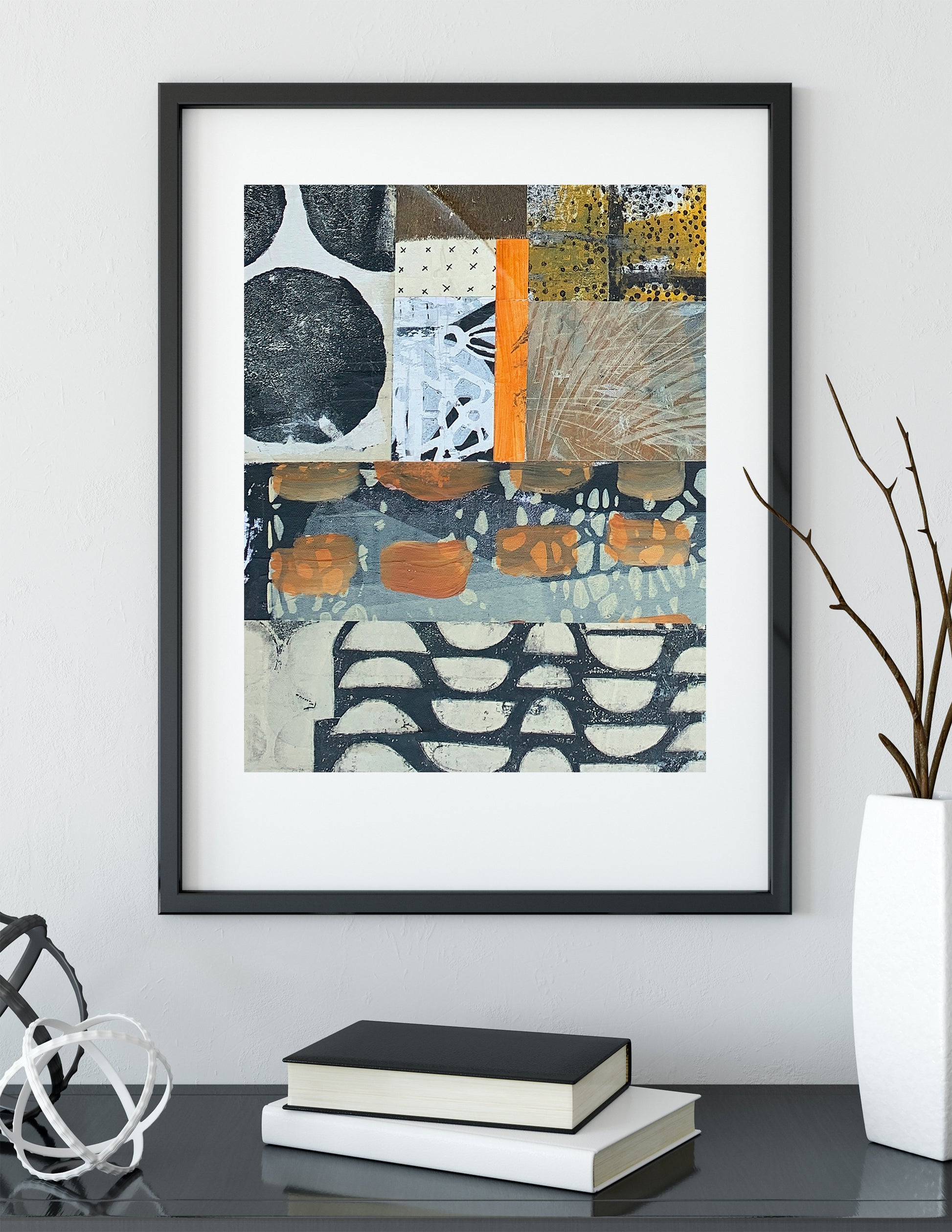 Framed abstract wall art hanging on a light gray wall above a dark table with stacked books and a white vase with branches. The artwork has a black frame, white mat, and an abstract patchwork design with navy, gray, beige, black, white, and orange blocks.