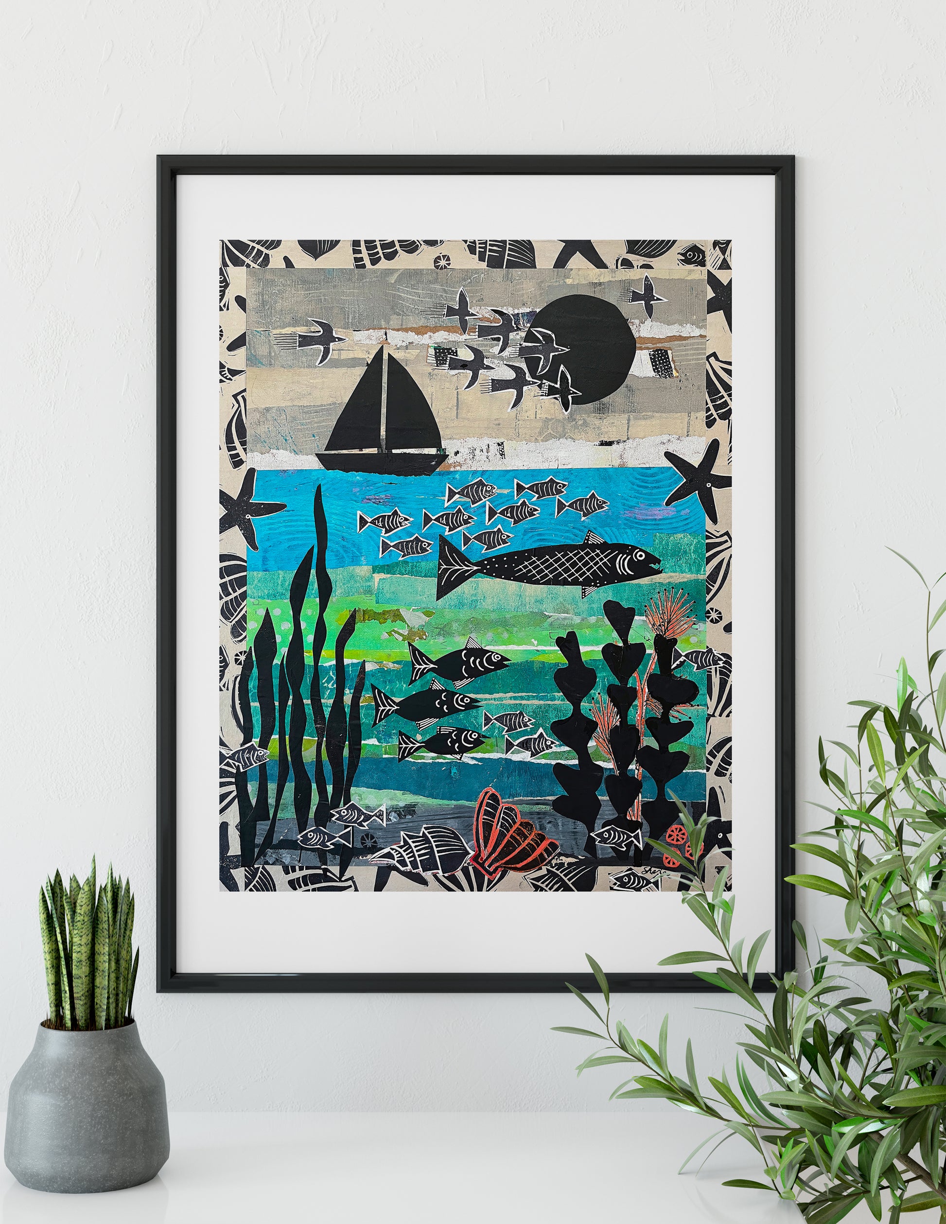 A framed art print featuring a stylized underwater seascape with fish, a sailboat, starfish, and kelp, displayed on a white wall above a shelf with a small potted plant and green foliage.