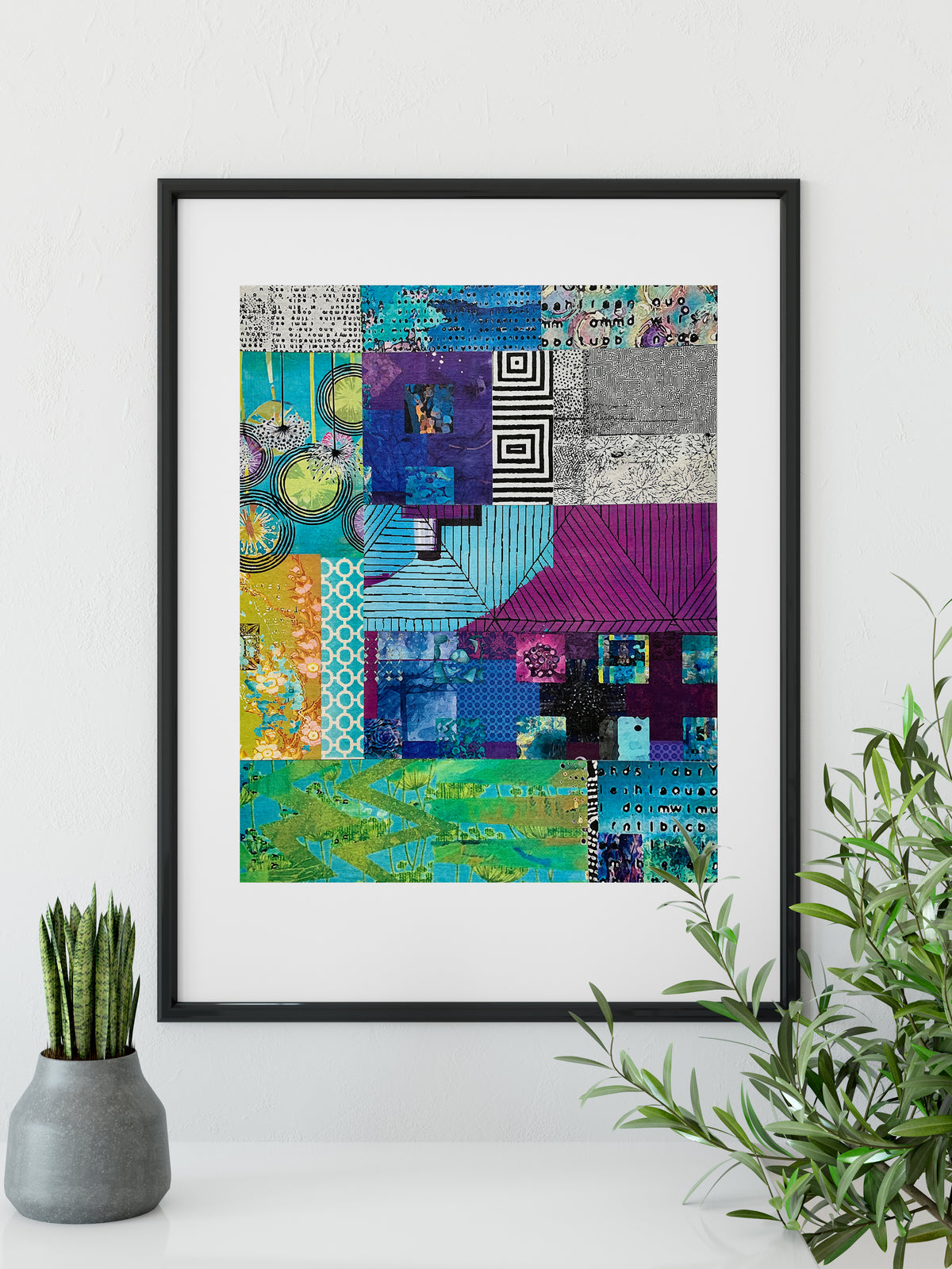 A framed abstract art print featuring a vibrant patchwork design with blues, purples, greens, and grays, styled on a white surface with a small potted succulent and a green plant in the foreground.