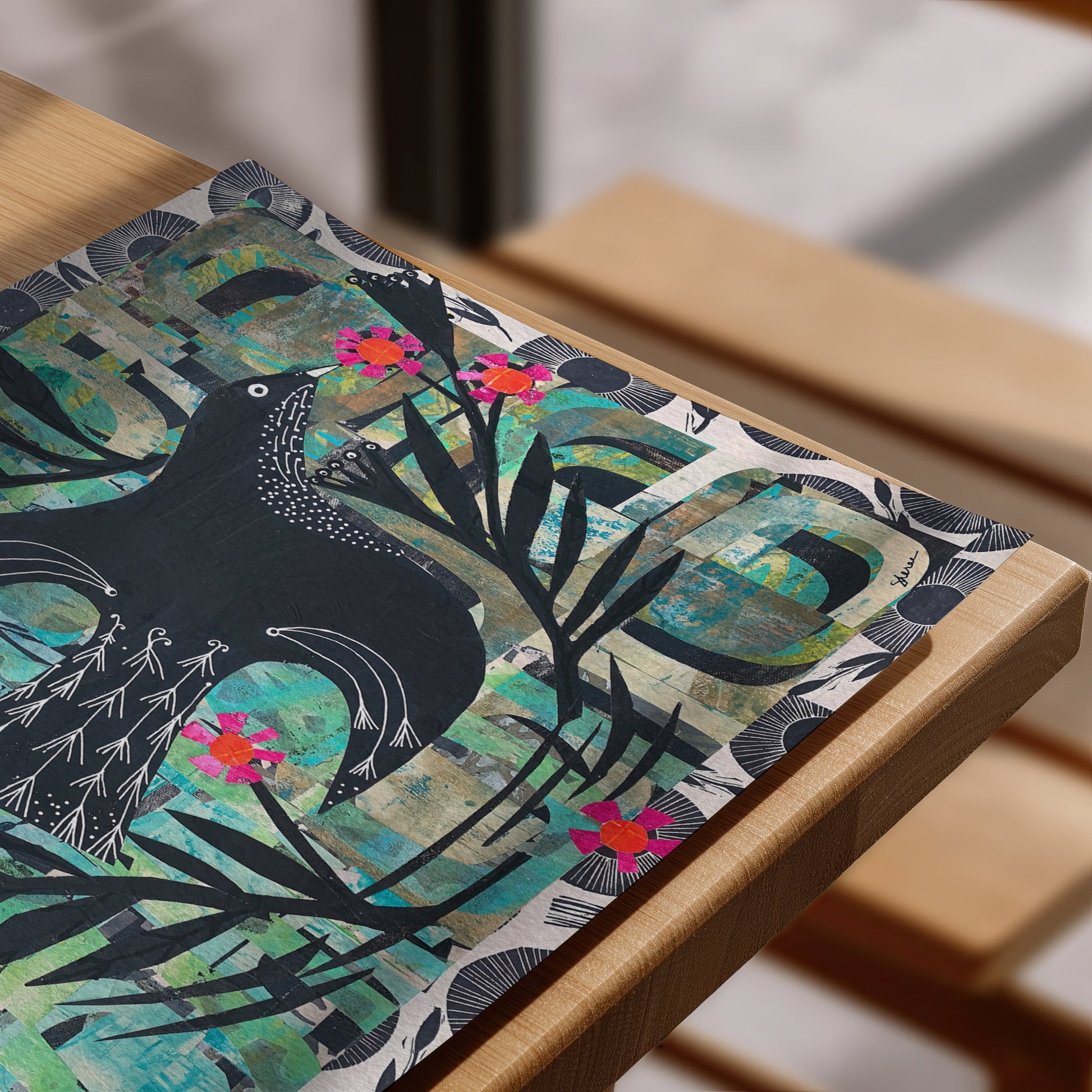 A rectangular art print or placemat featuring a stylized black bird, teal and green textured background, and bright pink flowers, displayed on a light wood table.