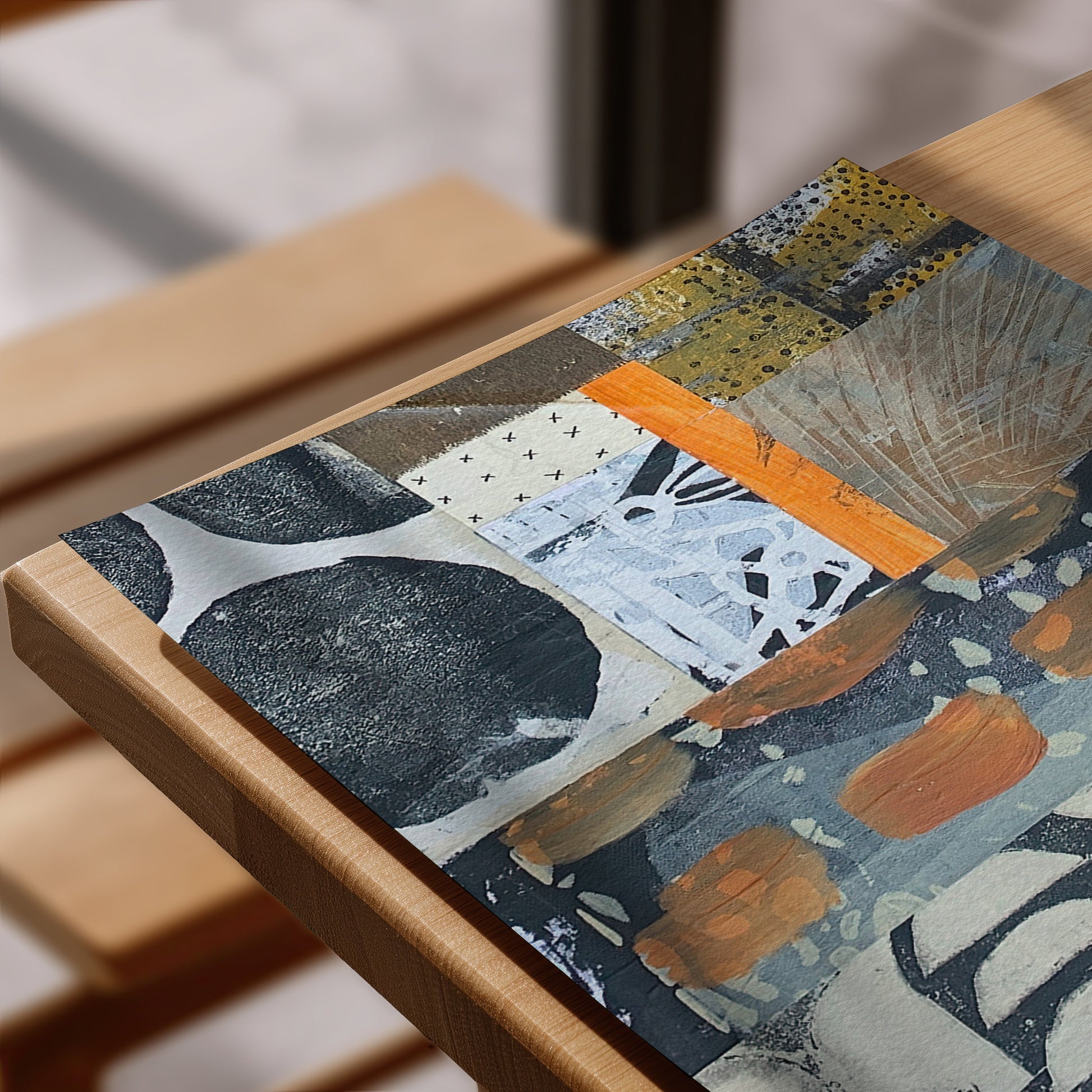An abstract patchwork art print featuring black, white, orange, and yellow patterns, displayed on a light wood tabletop.