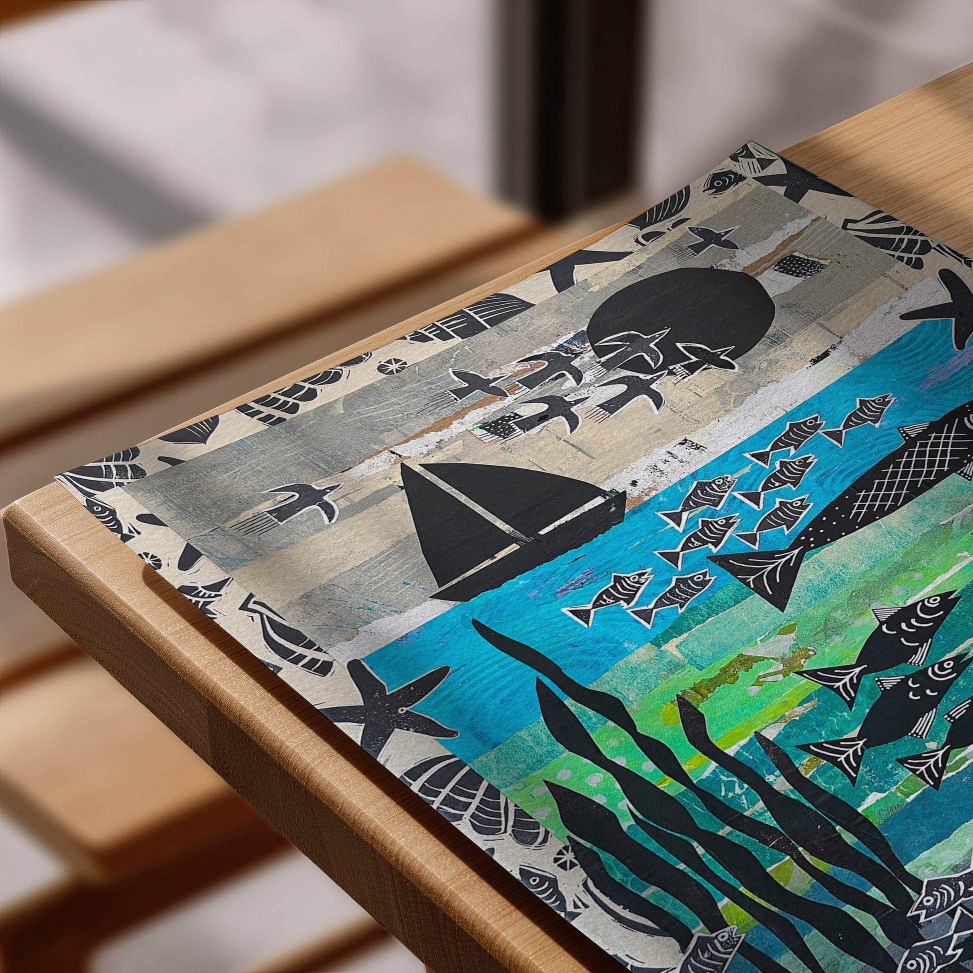 A square decorative placemat featuring an artistic illustration of an ocean scene, displayed on a light wood table. The design includes black sailboats, fish, starfish, and kelp on a textured background of gray, beige, and bright blue.