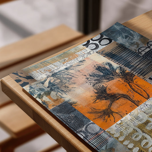 A rectangular art print featuring an abstract collage design with orange, gray, black, and beige tones, resting on a light wood frame in a softly blurred indoor setting.