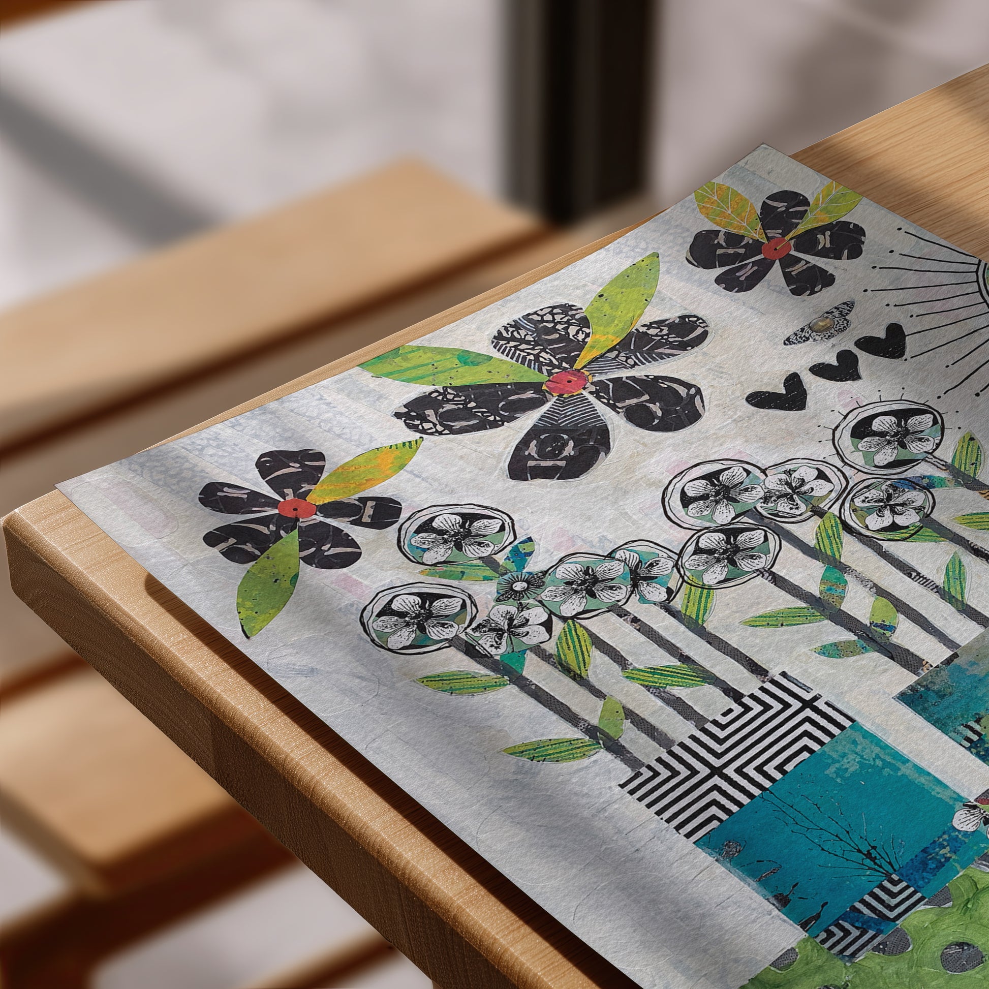 A close-up of a rectangular printed artwork resting on a light wood  frame. The design features stylized black flowers with green and yellow leaves, teal floral motifs, black-and-white geometric patterns, and a turquoise section near the bottom right, all on a textured off-white background.