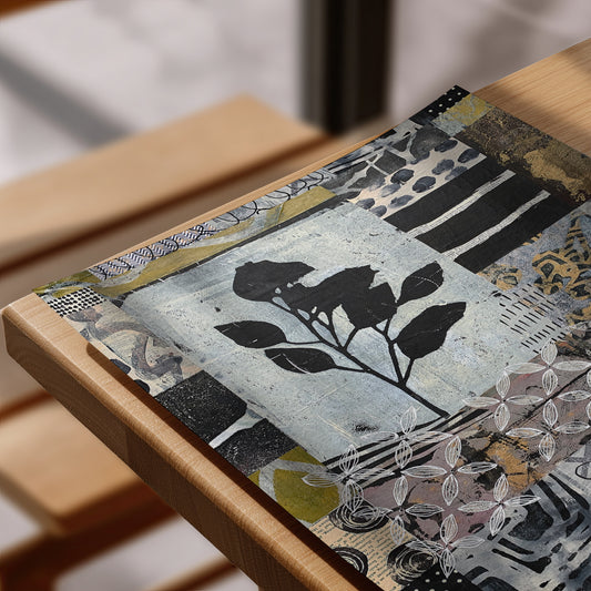 A rectangular art print or placemat featuring a mixed-media collage with black leaf silhouettes over a textured light blue-gray background, styled on a light wood table.