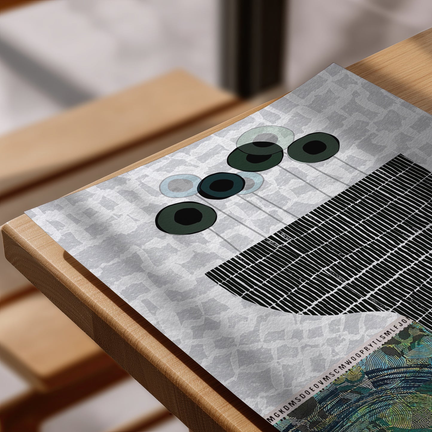 A close-up of a printed art print or poster laid on a wooden table. The artwork features abstract circular forms in dark green, black, and pale blue over a light gray patterned background, with a black grid-like section and a small colorful landscape-like area in the lower right.