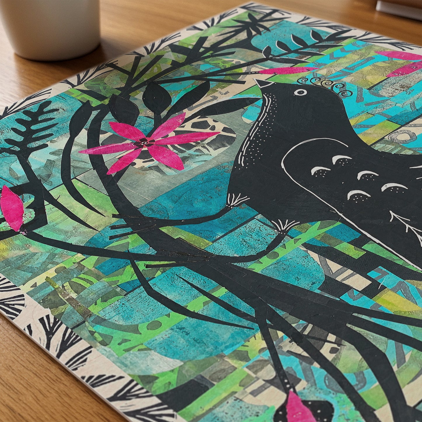 A square art print or featuring a stylized black bird perched on branches with bright pink flowers over a collage background of turquoise, green, beige, and yellow tones, photographed on a wooden surface.