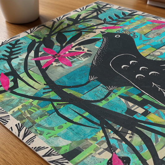 A square art print or featuring a stylized black bird perched on branches with bright pink flowers over a collage background of turquoise, green, beige, and yellow tones, photographed on a wooden surface.