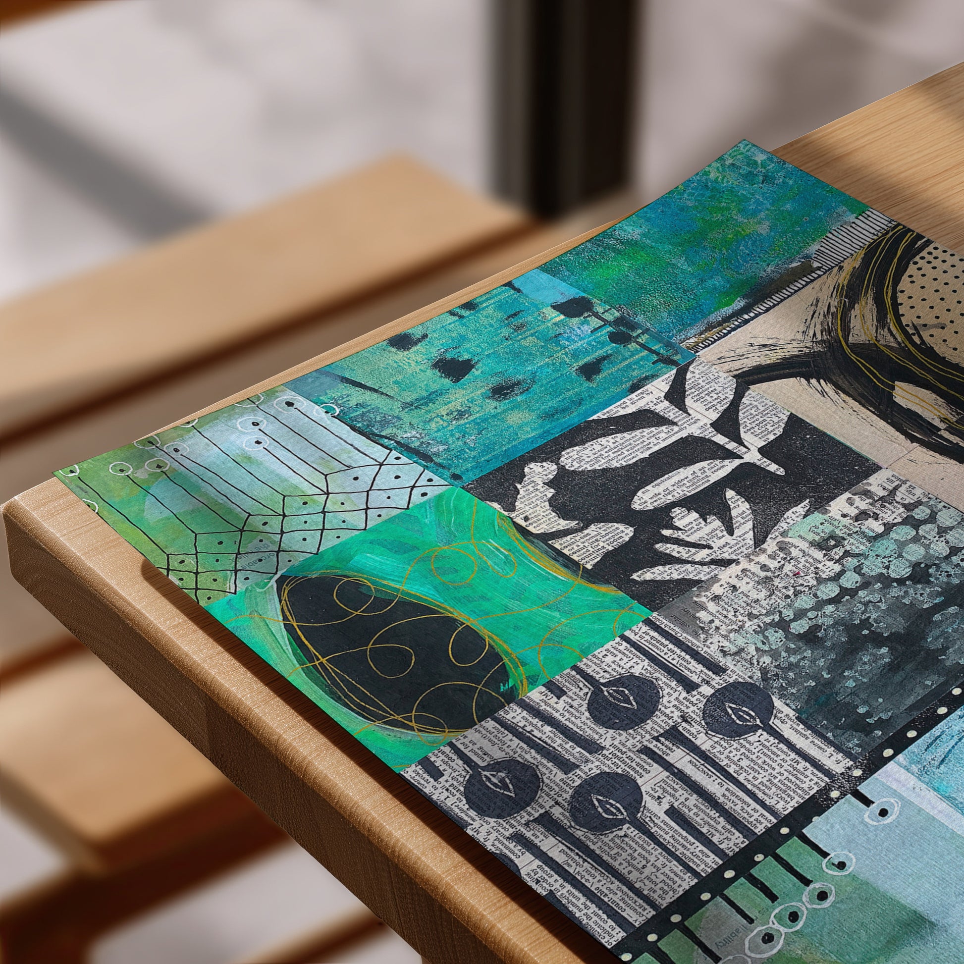A rectangular mixed-media art print or placemat featuring an abstract collage of teal, green, black, white, and gray textures, shown resting on a light wood table.