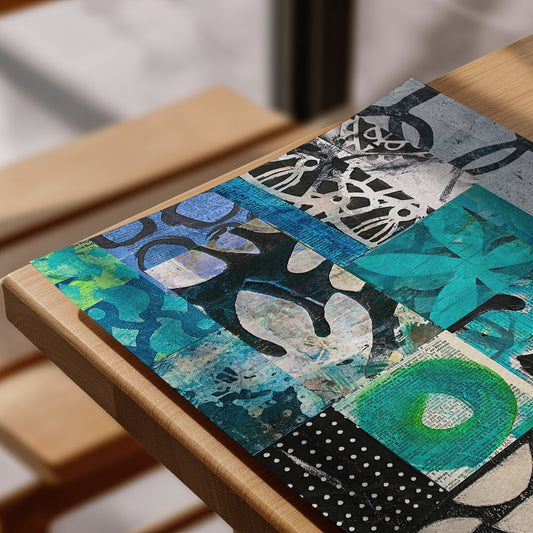 A square abstract art print with a patchwork design in teal, blue, black, white, and green, displayed on a light wood table.