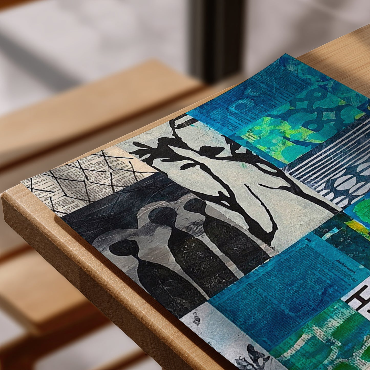 A rectangular mixed-media collage art print featuring blue, black, white, and green abstract patterns, displayed on a light wood table.