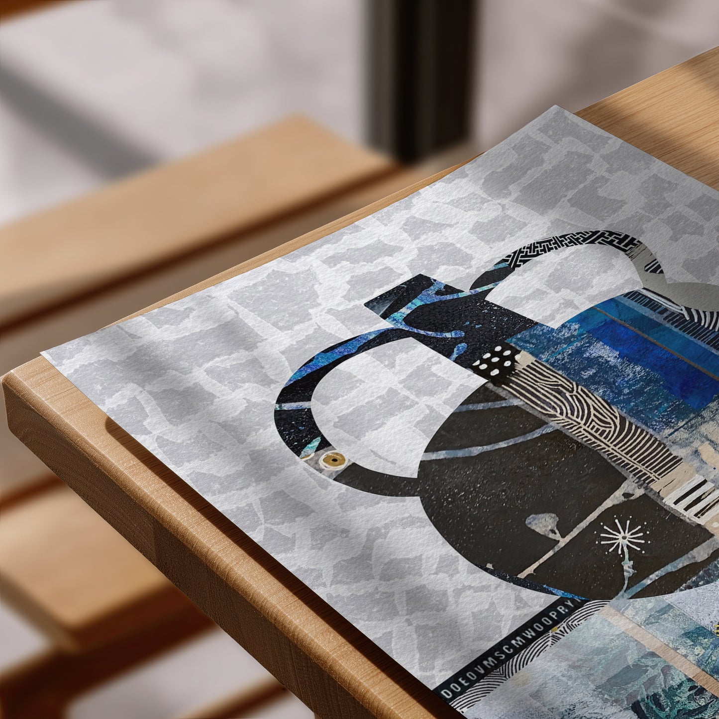 A close-up of a rectangular art print or poster laid on a wooden table, featuring an abstract collage design in black, blue, white, and gray.