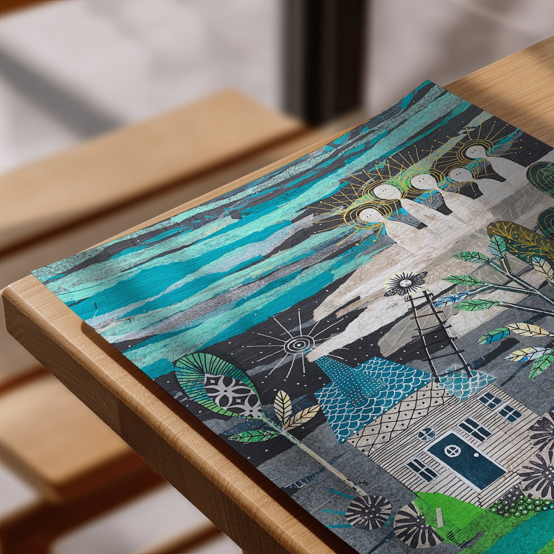 A close-up of a colorful art print featuring a whimsical house, trees, and abstract sky, resting on a light wood table.