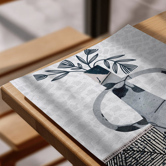 A close-up of a rectangular placemat or printed mat laid on a wooden table, featuring a minimalist navy illustration of a vase with leaves on a light gray patterned background.