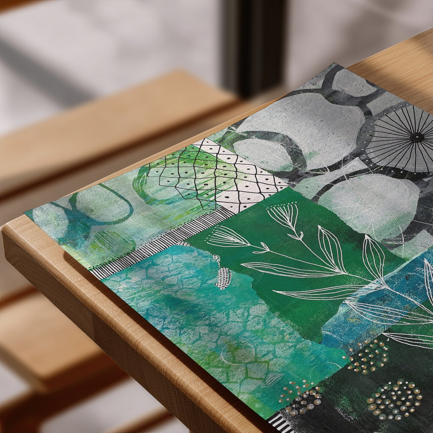 A square mixed-media art print featuring abstract botanical patterns in shades of green, teal, black, and white, displayed on a light wood table.
