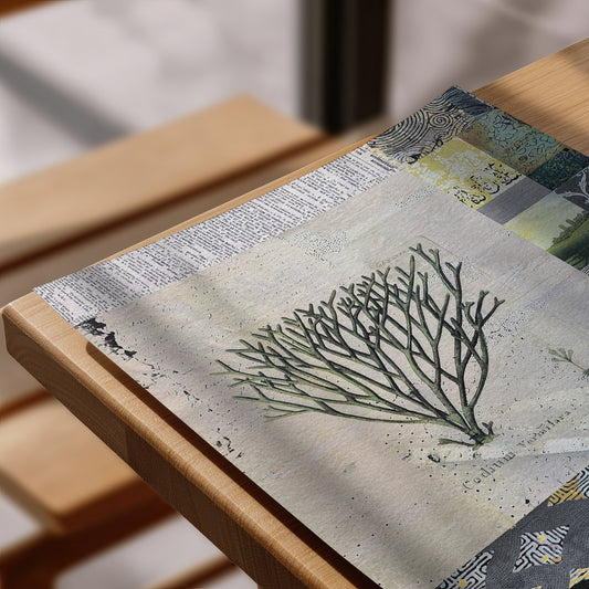 A close-up of a rectangular art print or placemat featuring a green coral-like branch pattern over a textured beige background with newspaper print and abstract blue-gray accents, resting on a light wood table.