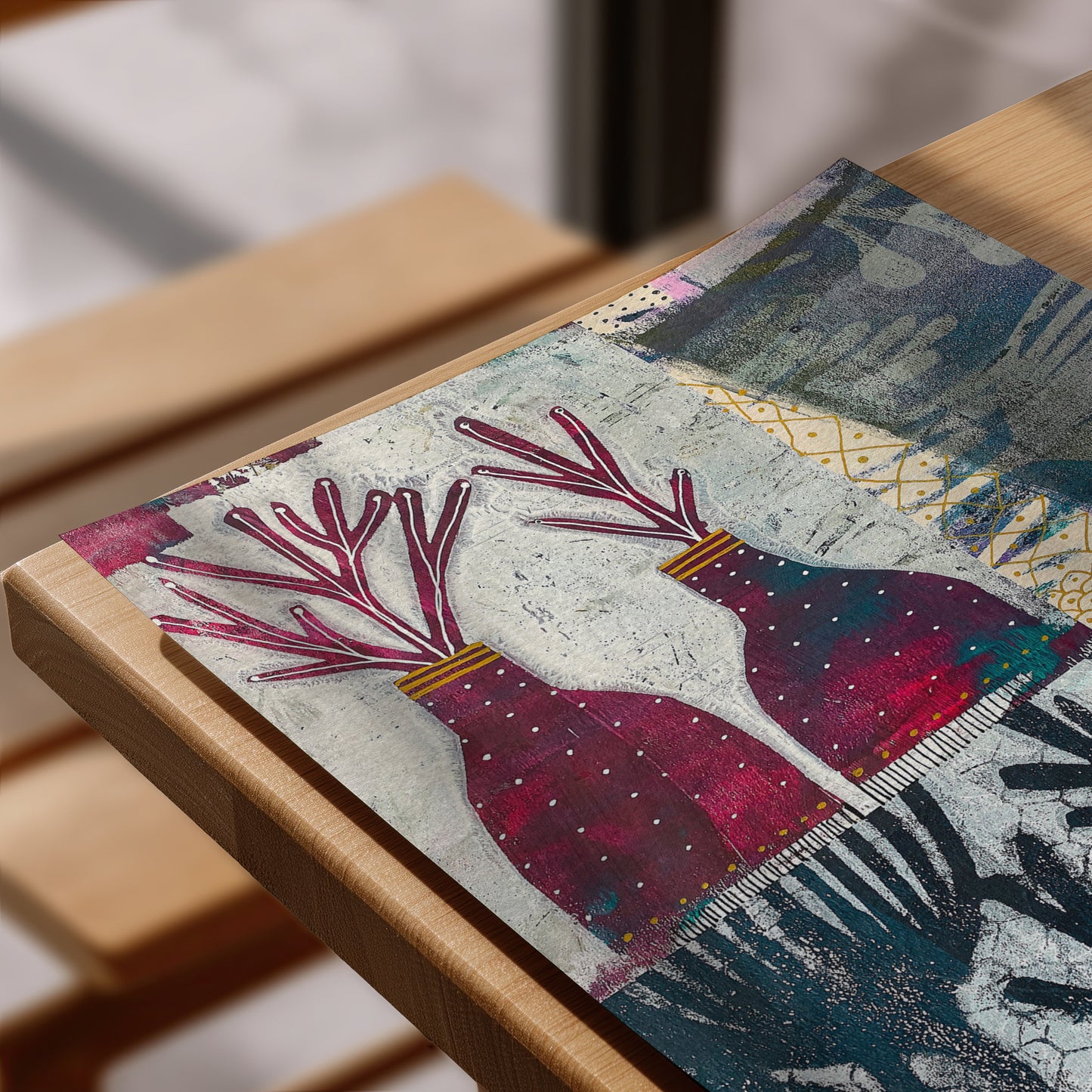 A close-up of a rectangular art print  laid on a light wood table. The artwork features two stylized deer heads with branching antlers in deep pink and burgundy, set against a textured off-white background with blue-gray and gold abstract patterns.