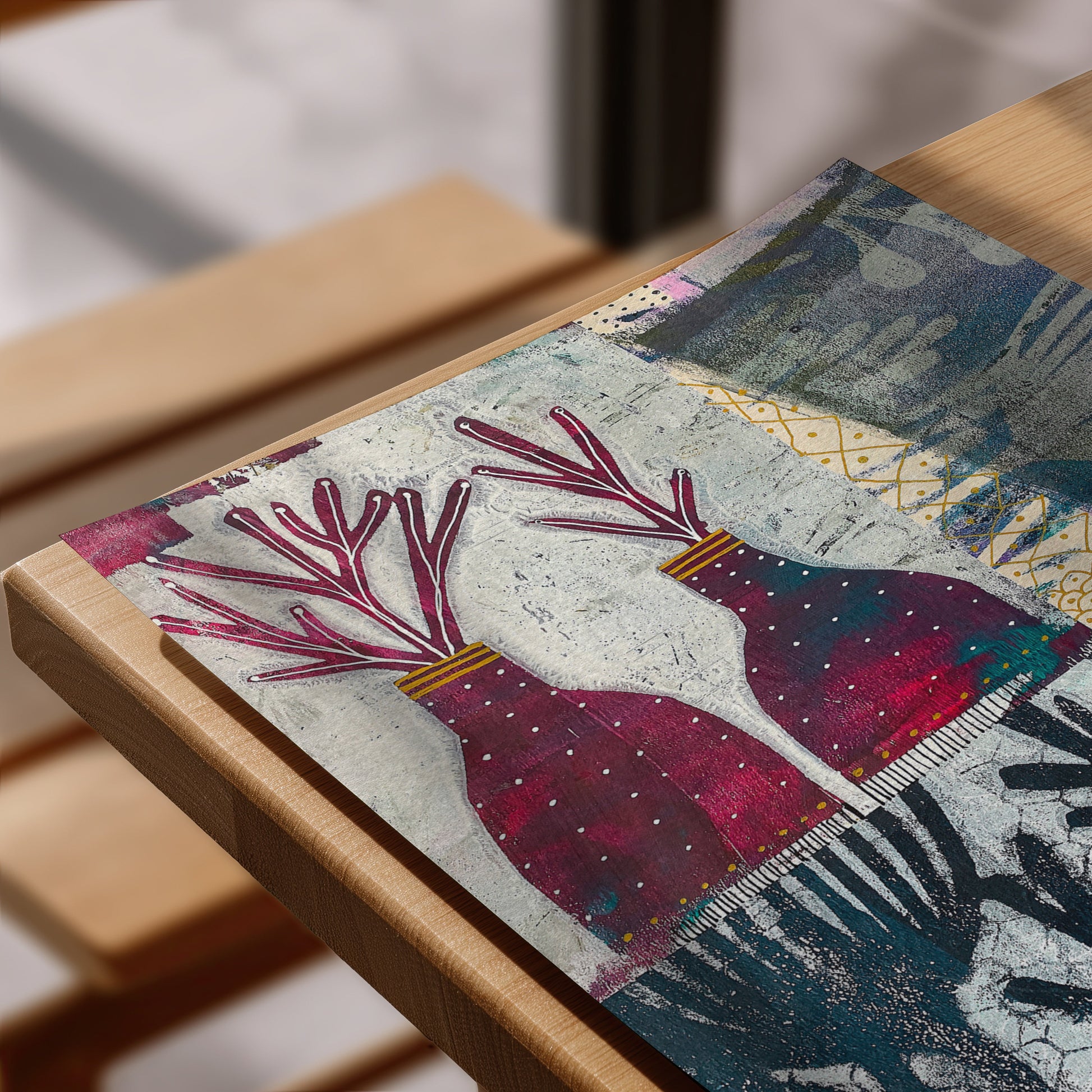 A close-up of a rectangular art print  laid on a light wood table. The artwork features two stylized deer heads with branching antlers in deep pink and burgundy, set against a textured off-white background with blue-gray and gold abstract patterns.