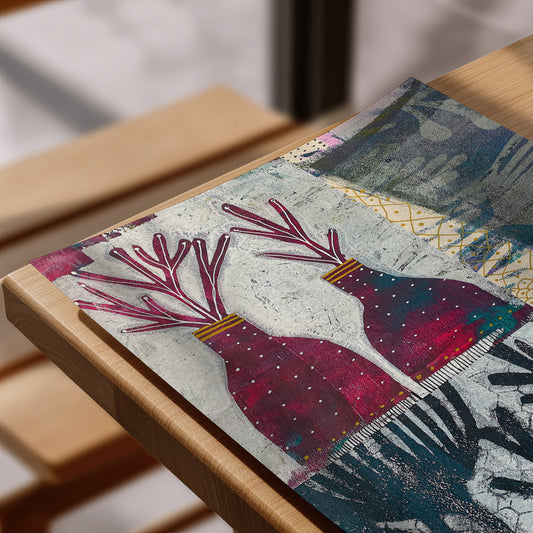 A close-up of a rectangular art print  laid on a light wood table. The artwork features two stylized deer heads with branching antlers in deep pink and burgundy, set against a textured off-white background with blue-gray and gold abstract patterns.