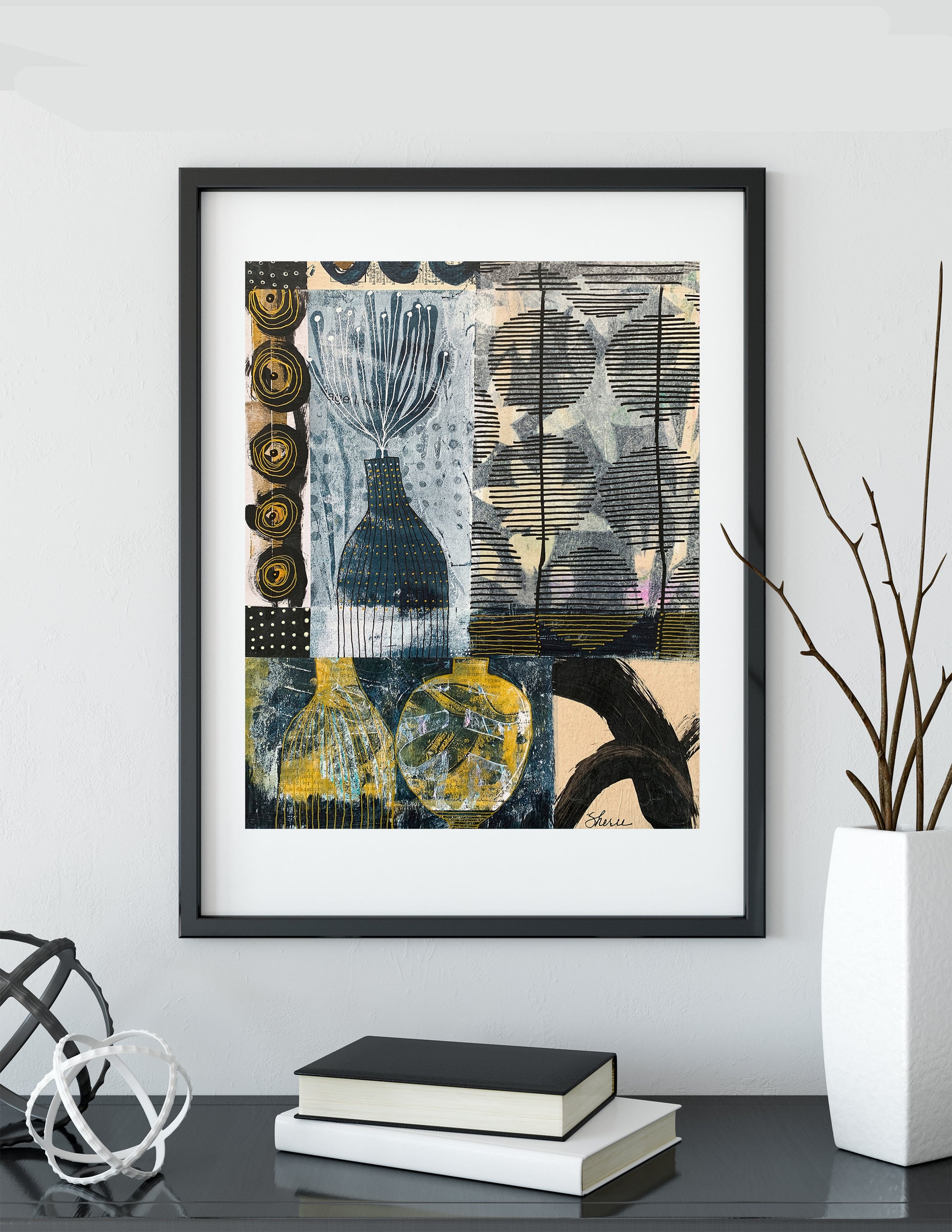 A framed abstract art print featuring a collage of black, gray, blue, and yellow patterns, displayed on a shelf next to books and a vase with branches.