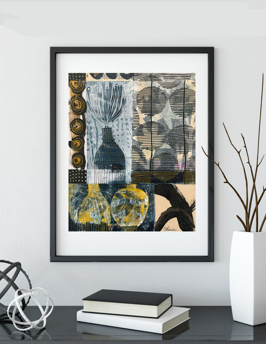 A framed abstract art print featuring a collage of black, gray, blue, and yellow patterns, displayed on a shelf next to books and a vase with branches.