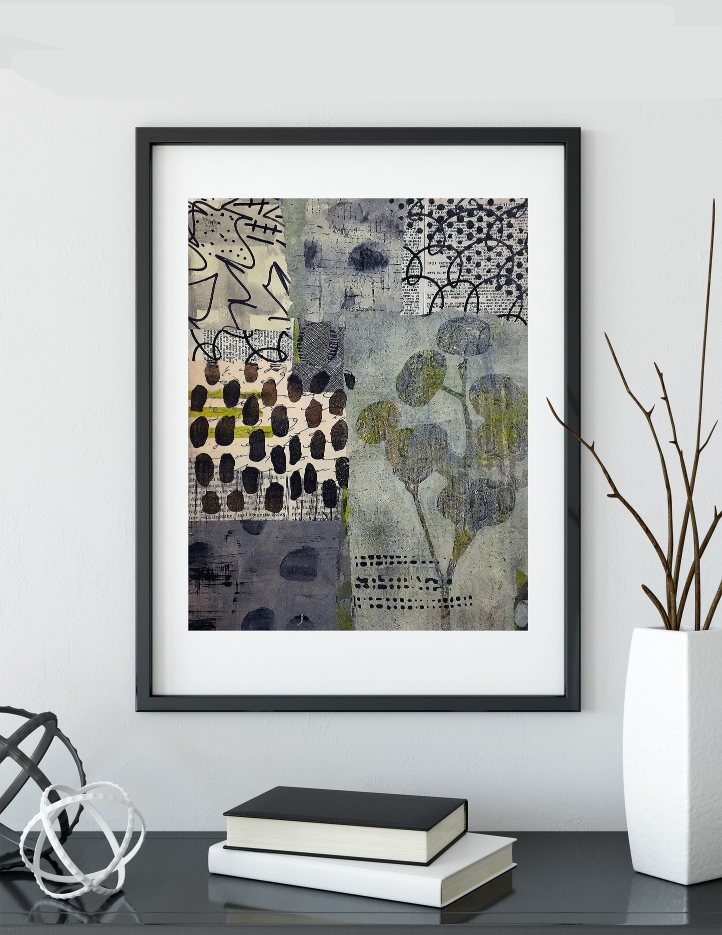 A framed abstract art print with a black frame and white mat, hanging on a light gray wall above a dark table with books, a white vase with branches, and decorative objects.
