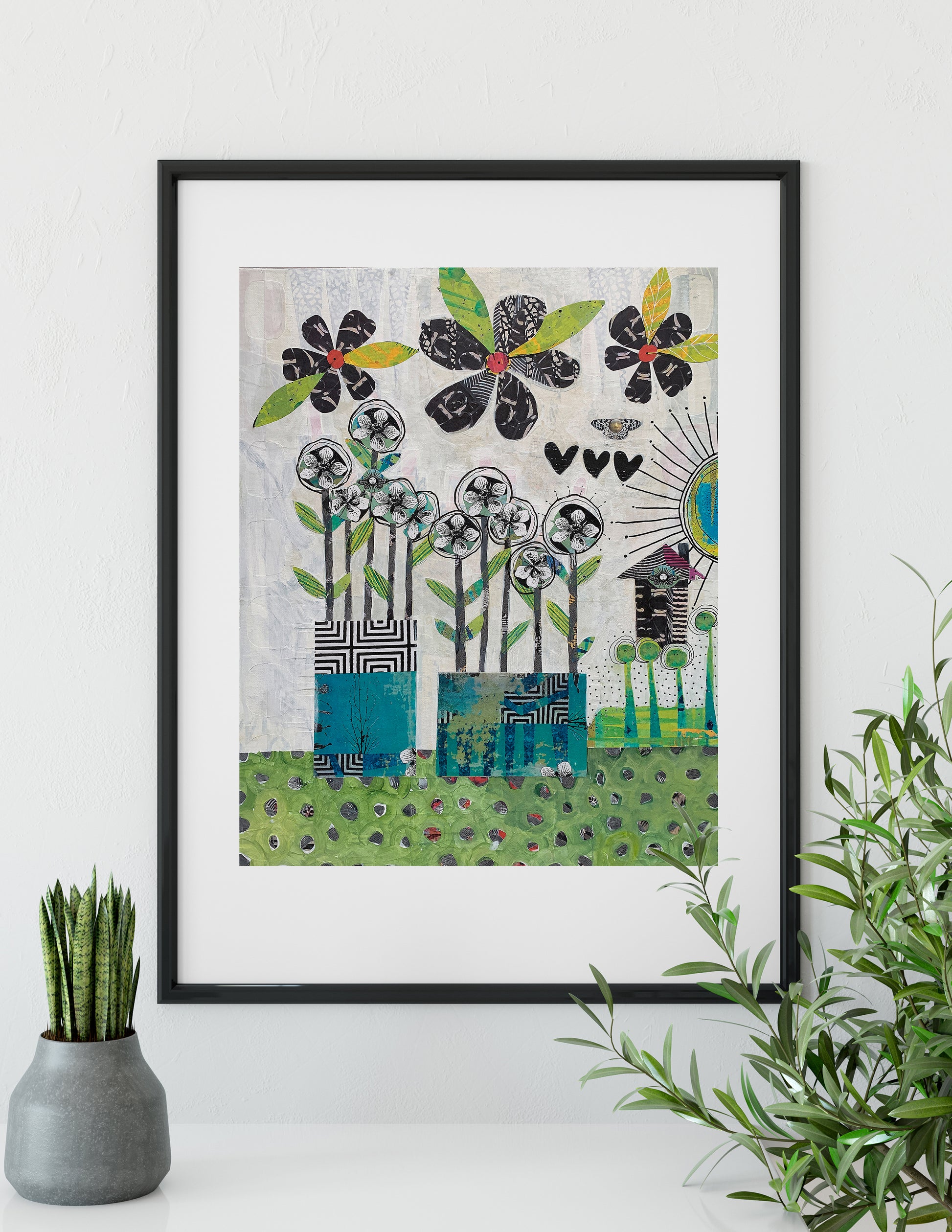 A framed art print featuring a whimsical, patterned illustration of flowers, birds, and a sun in black, green, blue, and beige tones, styled on a white wall above a small potted succulent and next to a green plant.
