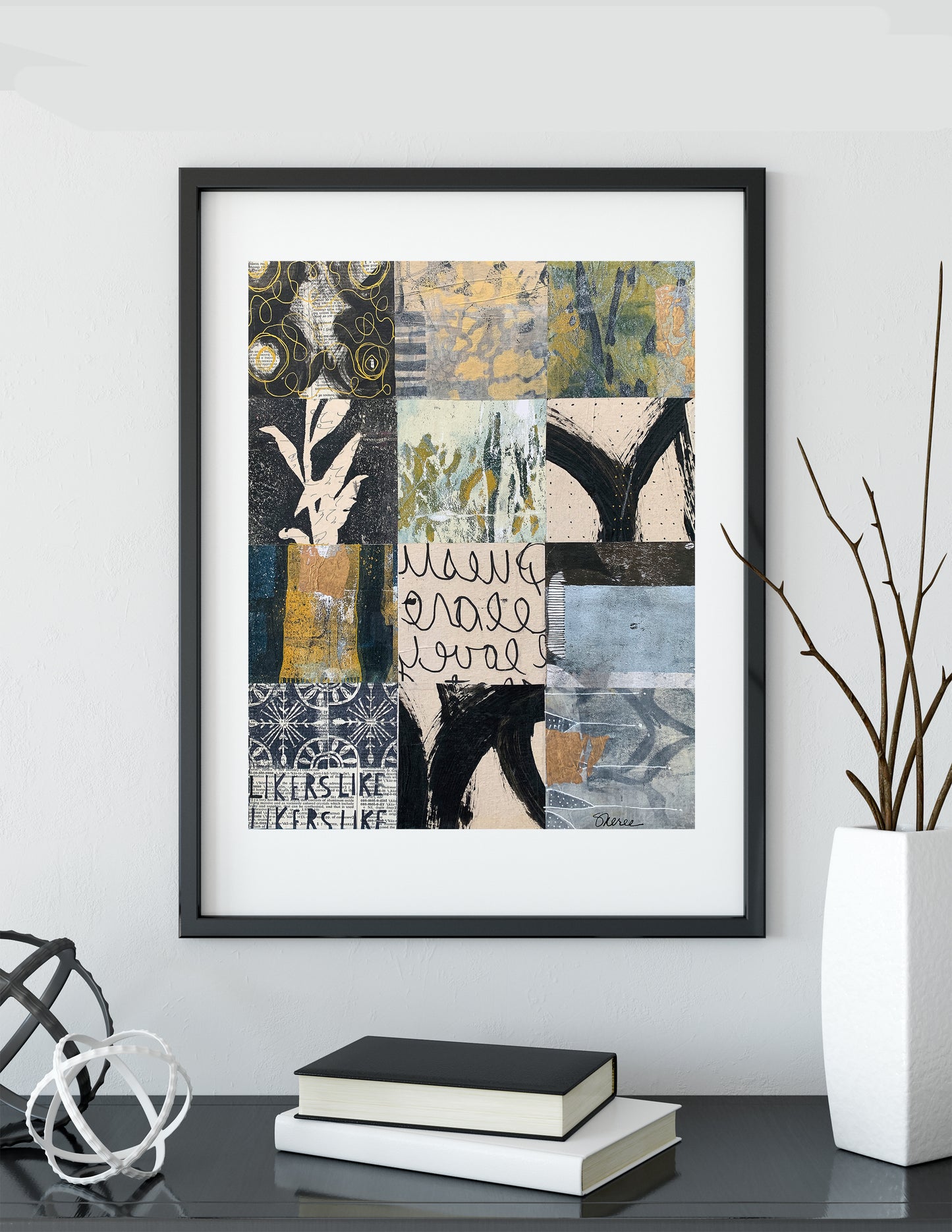 A framed abstract art print with a patchwork design in black, beige, blue, and green tones, displayed on a shelf in a modern room setting with books and a vase.