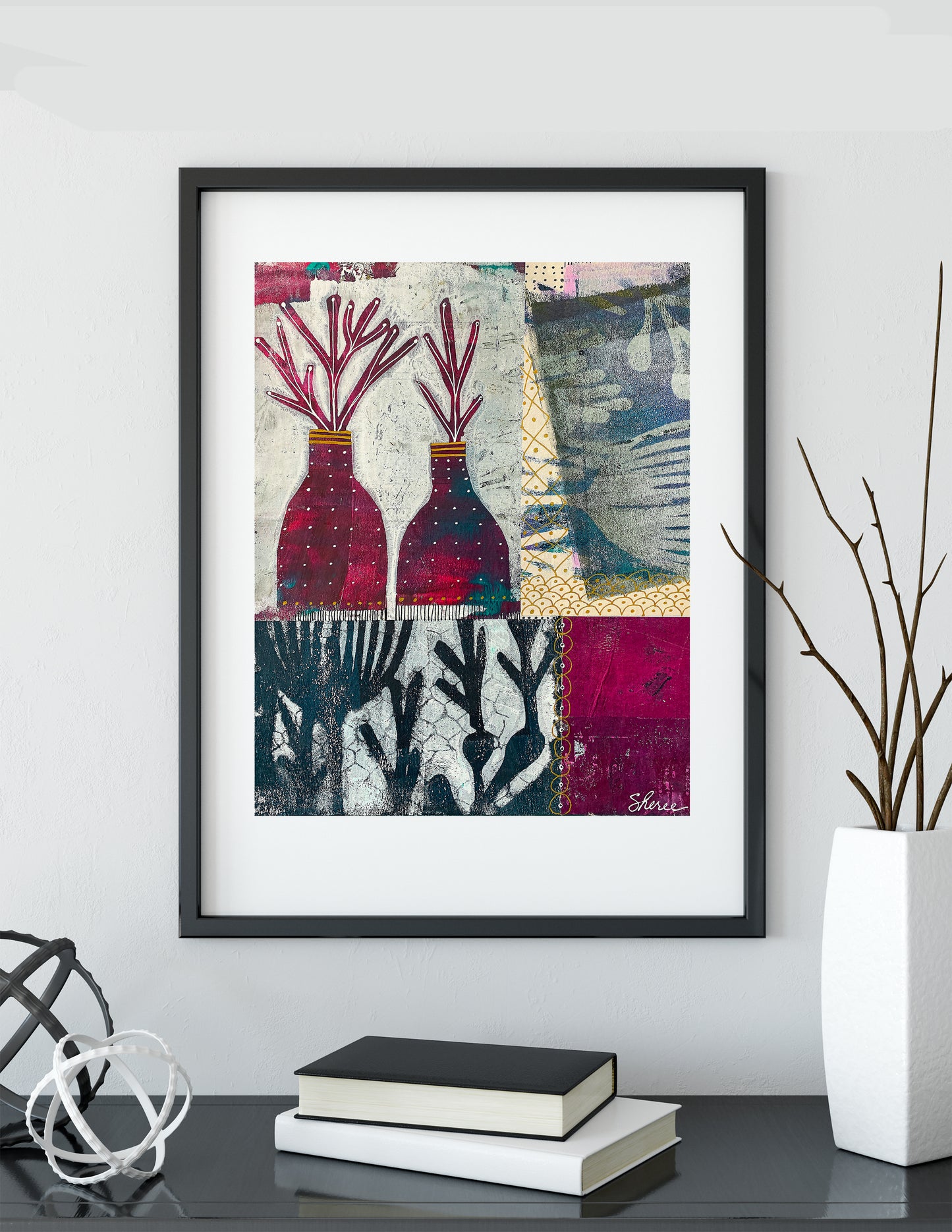 Framed abstract wall art hanging on a light gray wall above a dark tabletop. The artwork features two red and purple vase-like forms with branching stems on a patchwork background of cream, blue, and magenta.