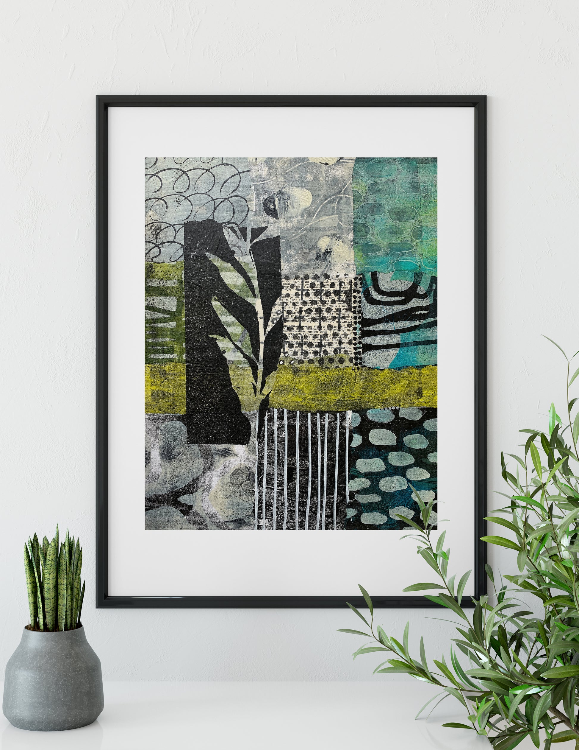 A framed abstract art print with a black frame and white mat, featuring a patchwork design in black, gray, teal, green, and yellow. The framed print is displayed on a white wall above a white surface, flanked by a small potted succulent on the left and a green plant on the right.