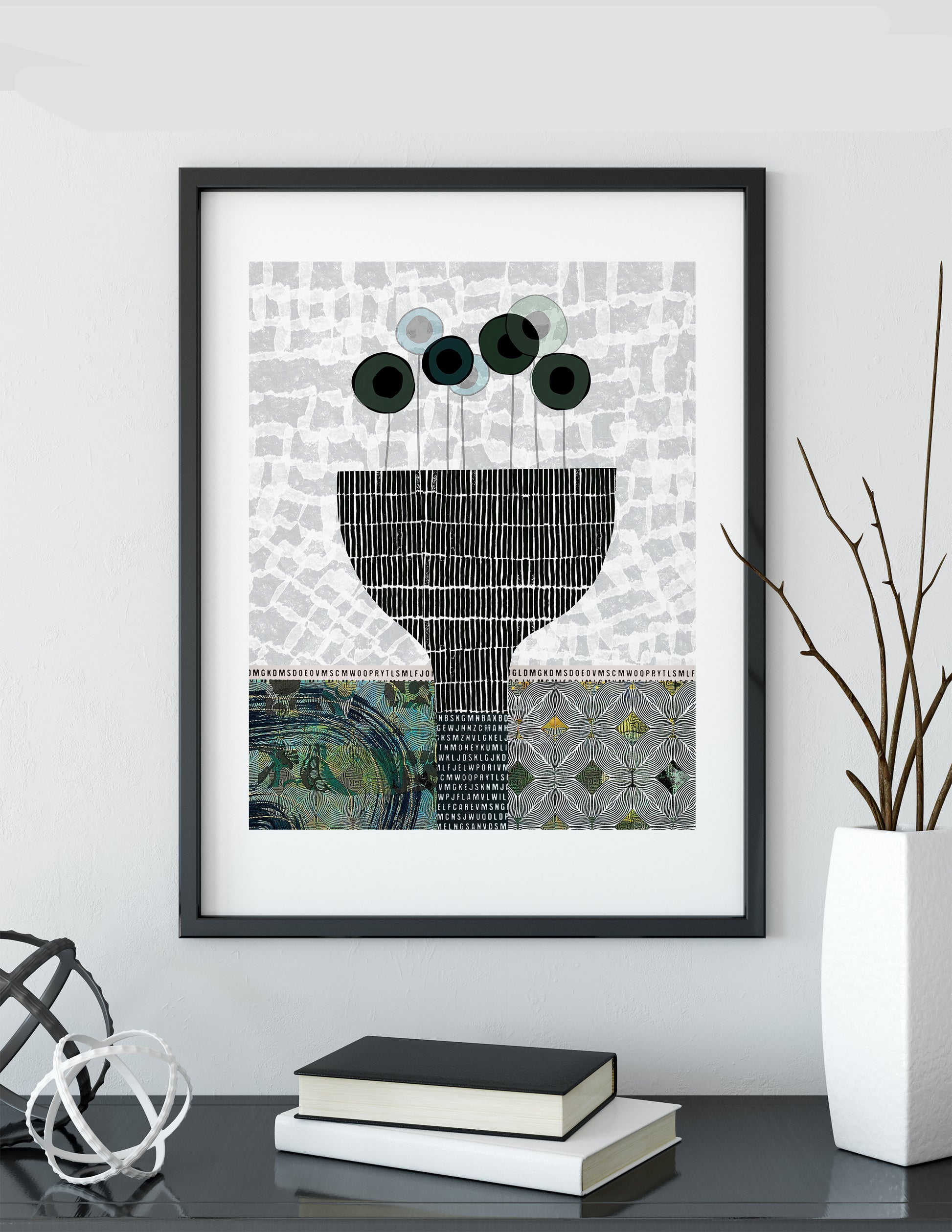 A framed abstract art print featuring a black and white patterned vase with dark green circular forms, displayed on a dark tabletop with books and a white vase in a styled interior setting.