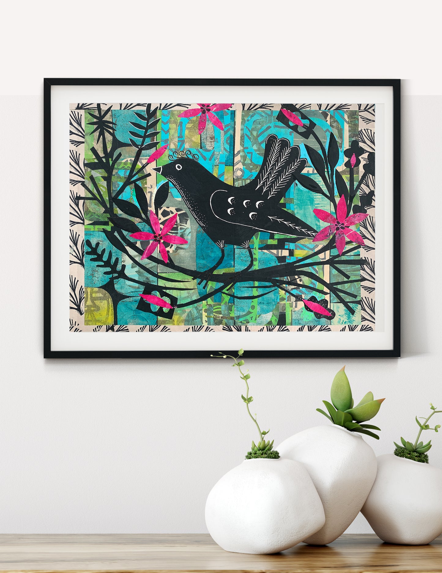 Framed wall art featuring a black bird perched on a branch with bright pink flowers and teal abstract background elements, displayed on a white wall above white ceramic planters on a wooden surface.