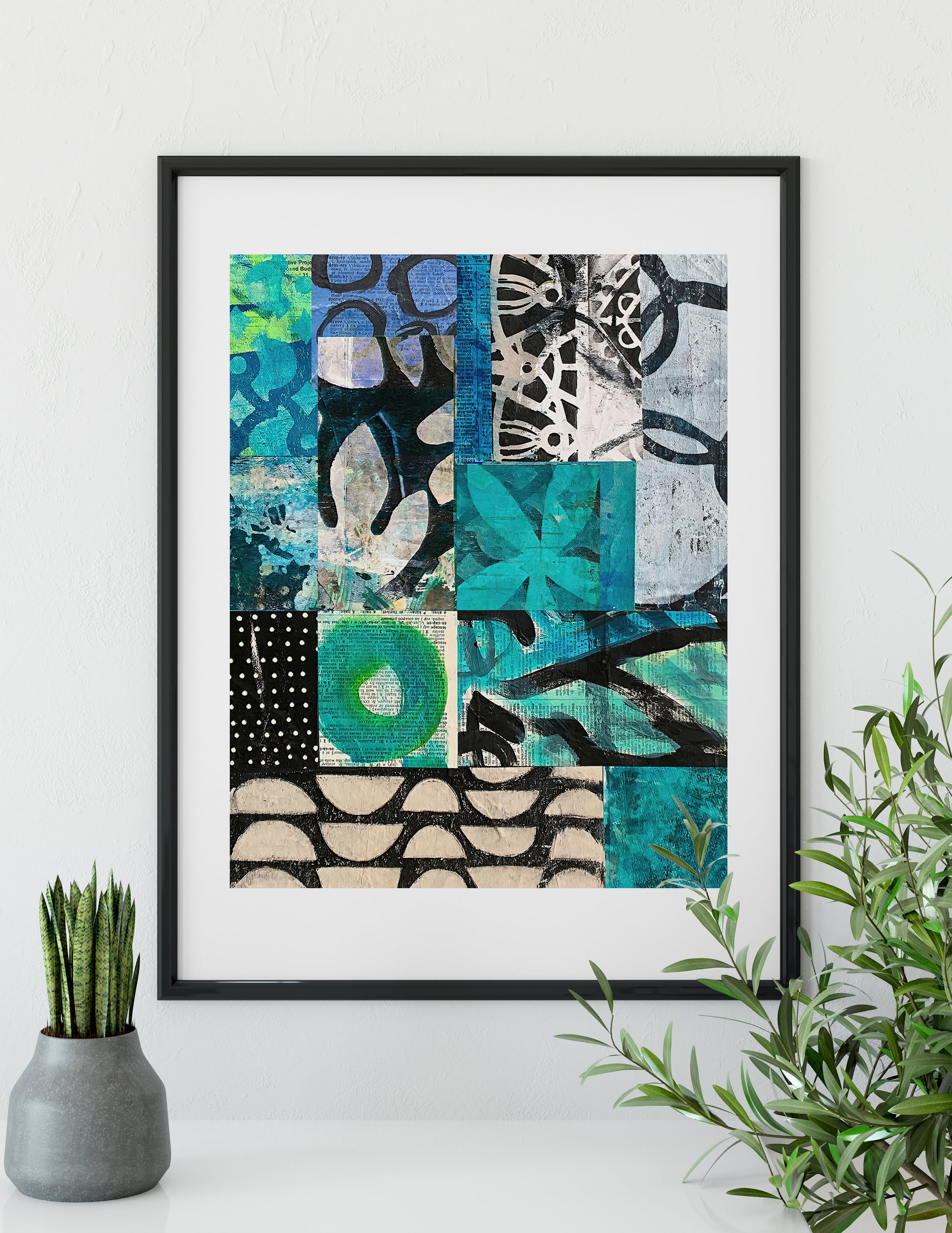A framed abstract art print featuring a patchwork of teal, green, black, white, and beige patterns, displayed on a white wall above a shelf with a small potted plant and a green leafy branch.