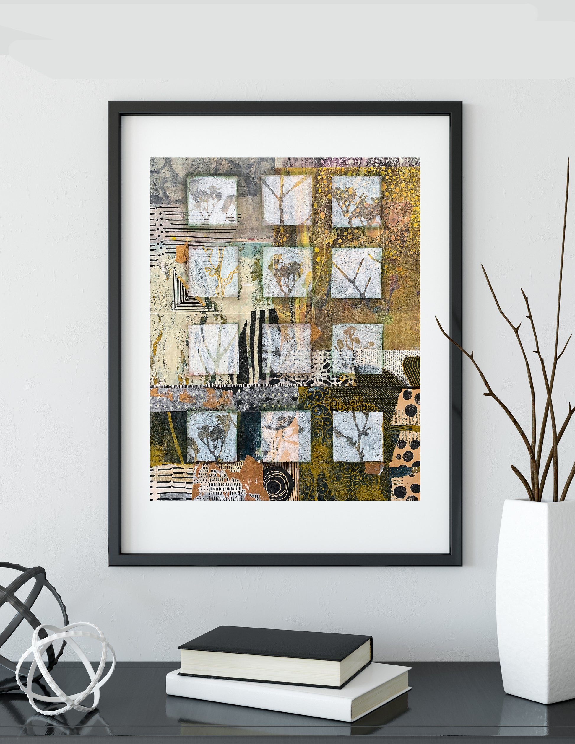 A framed abstract collage art print with a black frame and white mat, displayed on a dark table against a light gray wall, styled with books, a vase, and decorative objects.