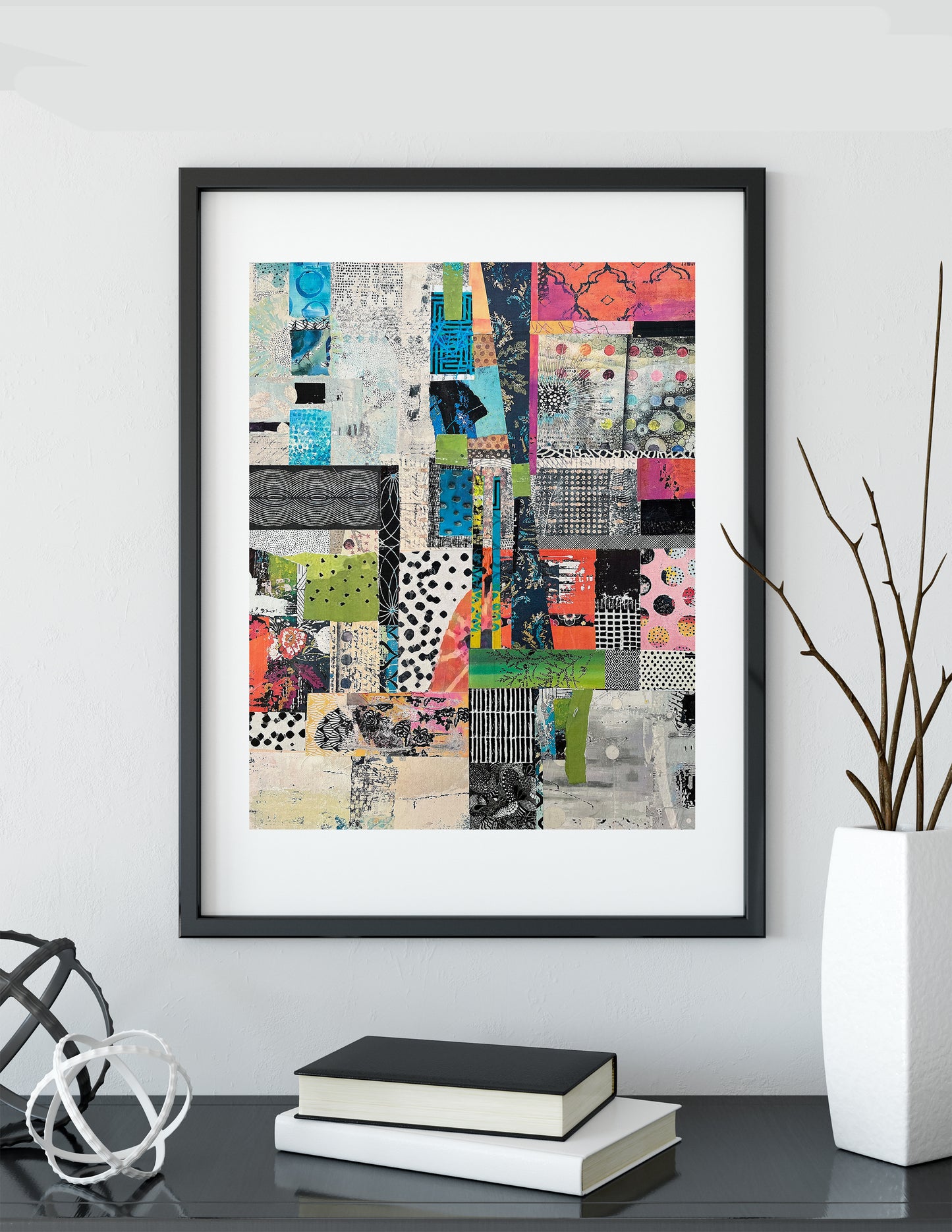 A framed abstract collage art print with a black frame and white mat, displayed on a shelf in a modern room setting with books and a vase.