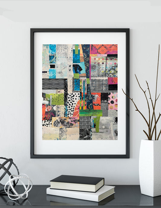 A framed abstract collage art print with a black frame and white mat, displayed on a shelf in a modern room setting with books and a vase.
