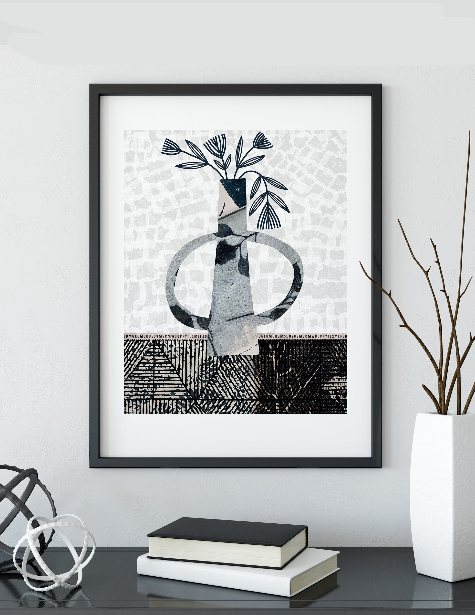 A framed abstract art print featuring a vase with leaves in black, white, and gray, displayed on a dark table with books and a vase of branches against a light gray wall.